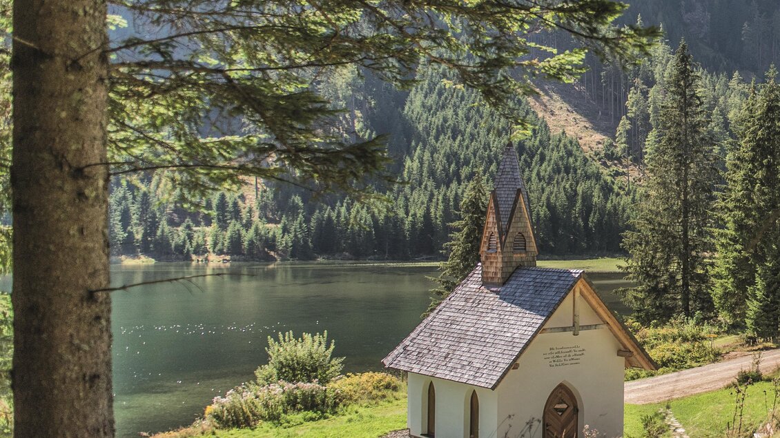 Eine kleine, malerische Kapelle steht in einer idyllischen Landschaft am See. Umgeben von hohen Bäumen und grünen Wiesen strahlt die Szene Ruhe und Frieden aus. | © Erlebnisregion Murtal