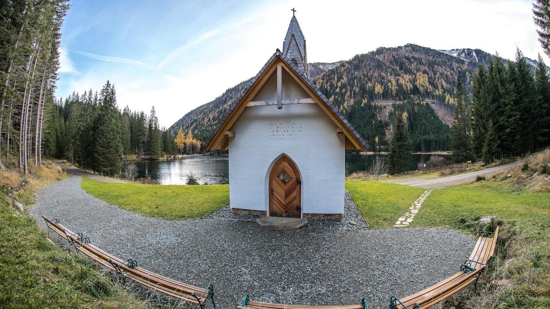 Eine kleine, weiße Kapelle am Ufer eines ruhigen Sees, umgeben von Bäumen und Bergen. Der Himmel ist klar und die Landschaft ist malerisch. | © Erlebnisregion Murtal