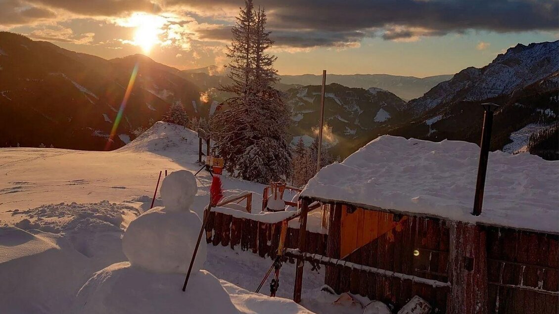 Eine verschneite Landschaft mit einem Sonnenuntergang im Hintergrund. Im Vordergrund sind eine Hütte und ein Schneemann zu sehen. | © Hirner