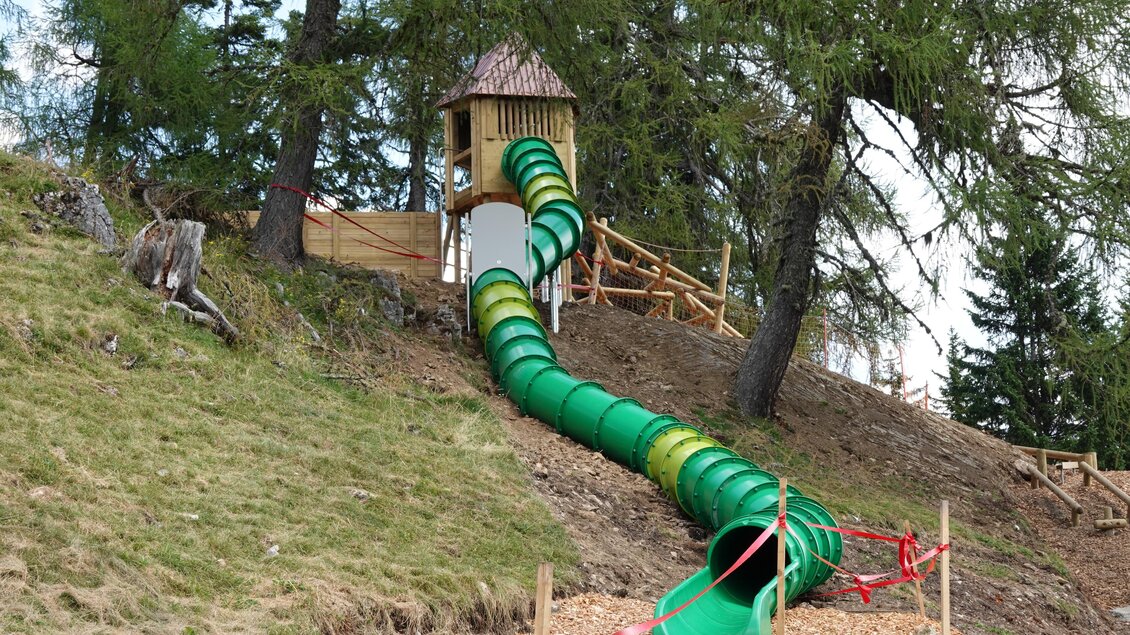 Eine lange, grüne Rutsche führt von einem Spielturm in die Höhe. Umgeben von Bäumen und einer Wiese, bietet sie viel Spaß für Kinder. | © Grebenzen Höhen