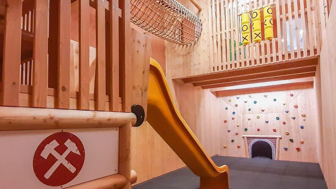 Ein Spielbereich mit einer Rutsche, Kletterwand und einem Netz zum Spielen. Die Wände sind aus Holz und es gibt bunten Klettersteine. | © JUFA Hotels