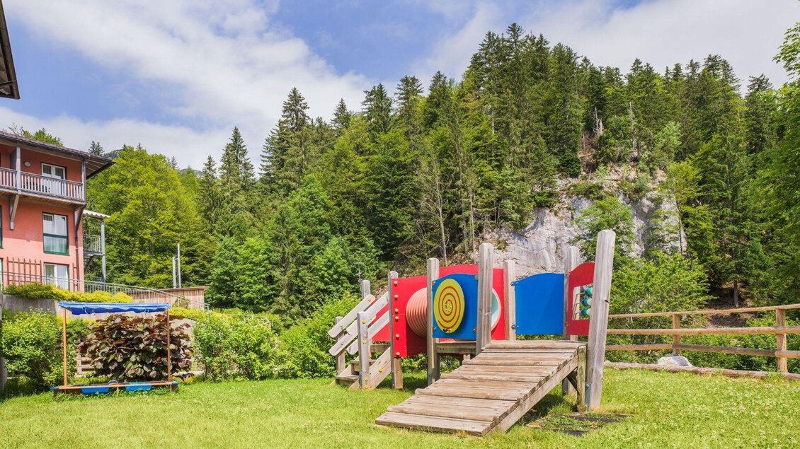 Ein Spielplatz in einer grünen Umgebung mit Holzspielgeräten und einer Rutsche. Im Hintergrund sind Bäume und ein Felsen zu sehen. | © JUFA Hotels