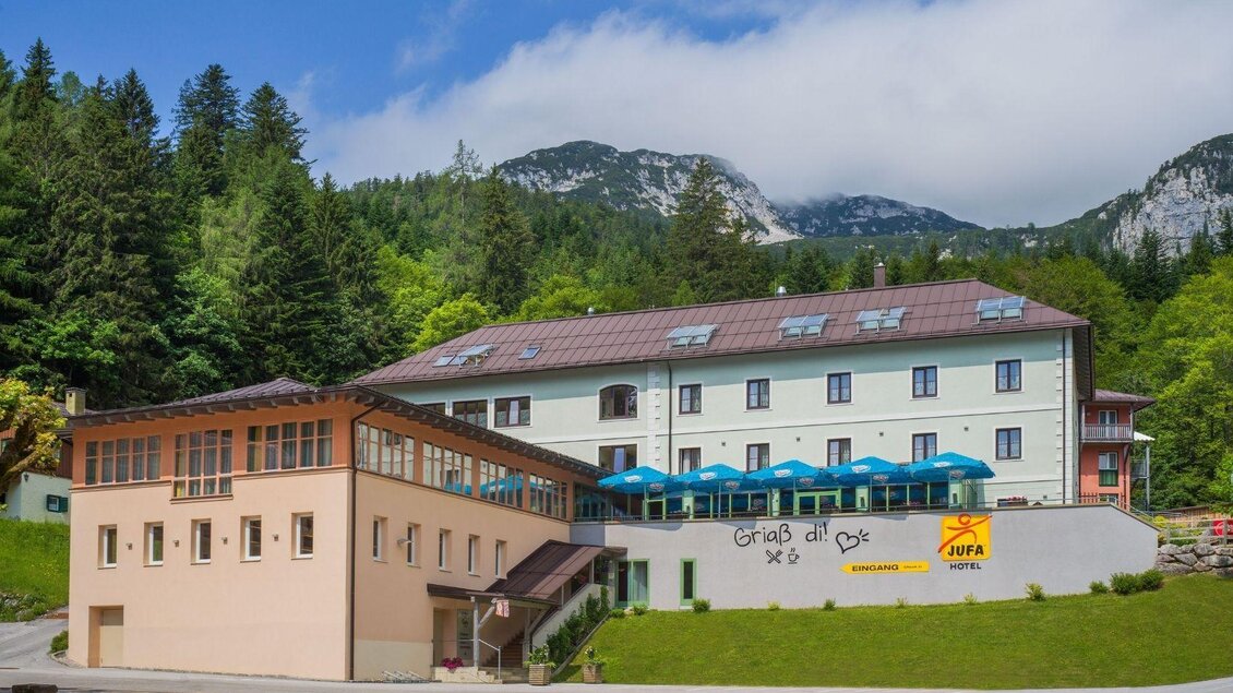 Ein modernes Hotel umgeben von Wald und Bergen. Auf der Vorderseite befinden sich Sonnenschirme und ein einladender Eingangsbereich. | © JUFA Hotels