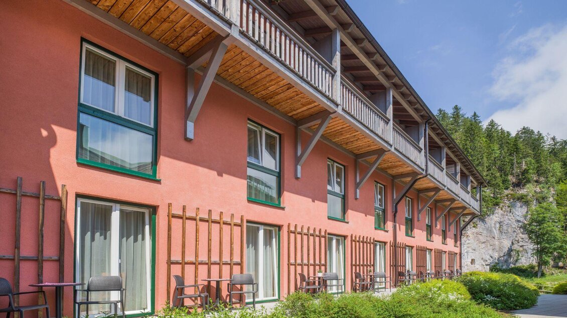 Ein modernes Gebäude mit roten Wänden und mehreren Fenstern. Draußen befinden sich Terrassen mit Stühlen und eine grüne Landschaft im Hintergrund. | © JUFA Hotels