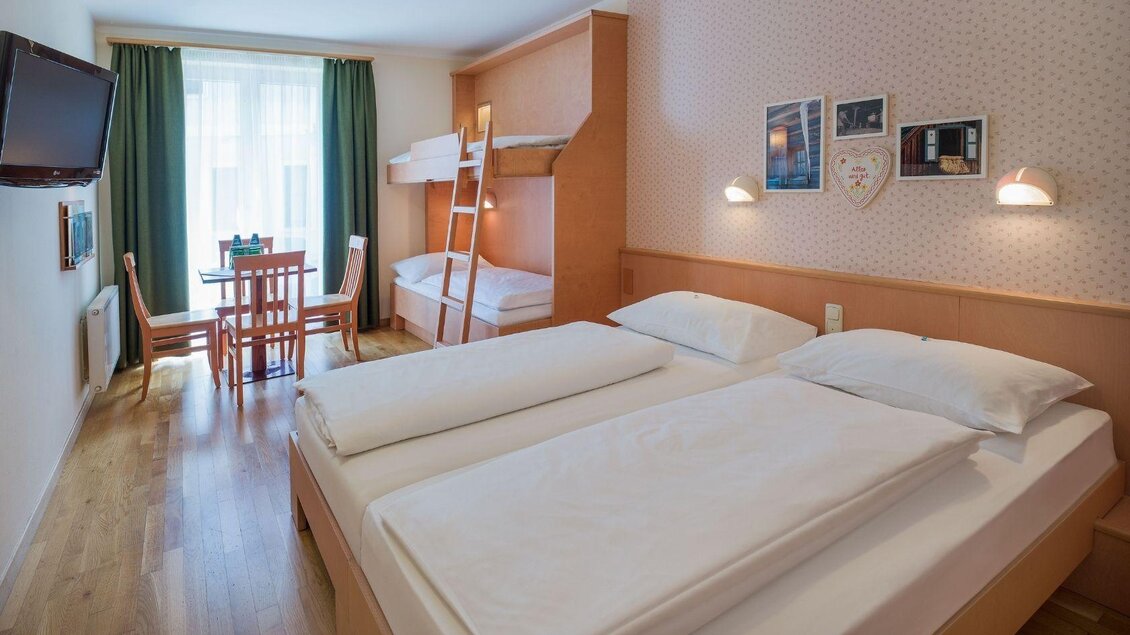Ein helles Hotelzimmer mit einem Doppelbett und einem Etagenbett. Ein Tisch mit Stühlen und ein Fernseher sind ebenfalls vorhanden. | © JUFA Hotels