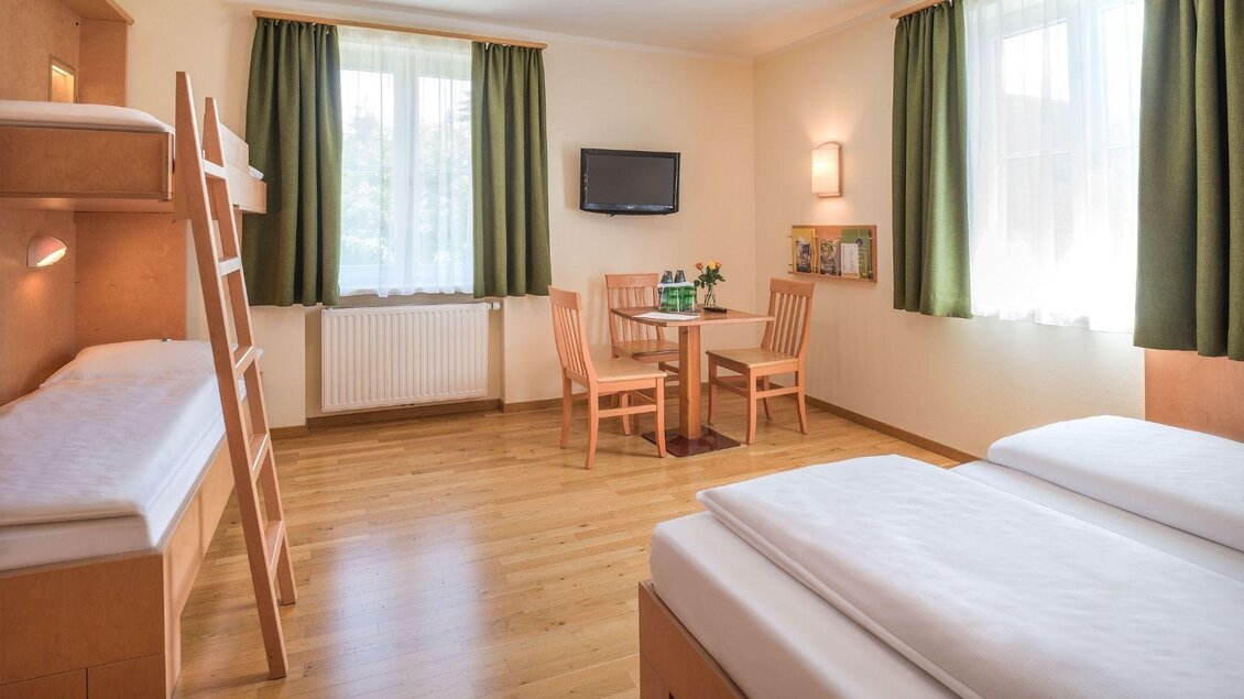 Ein helles Zimmer mit zwei Betten und einem Hochbett. Es gibt einen Tisch mit Stühlen und große Fenster mit grünen Vorhängen. | © JUFA Hotels
