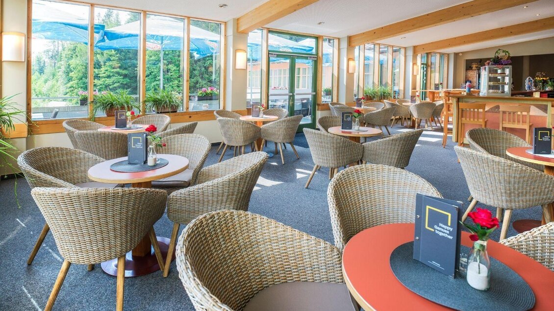 Ein modernes Café mit gemütlichen Rattan-Stühlen und großen Fenstern. Der Raum ist hell und einladend mit dekorativen Blumen auf den Tischen. | © JUFA Hotels