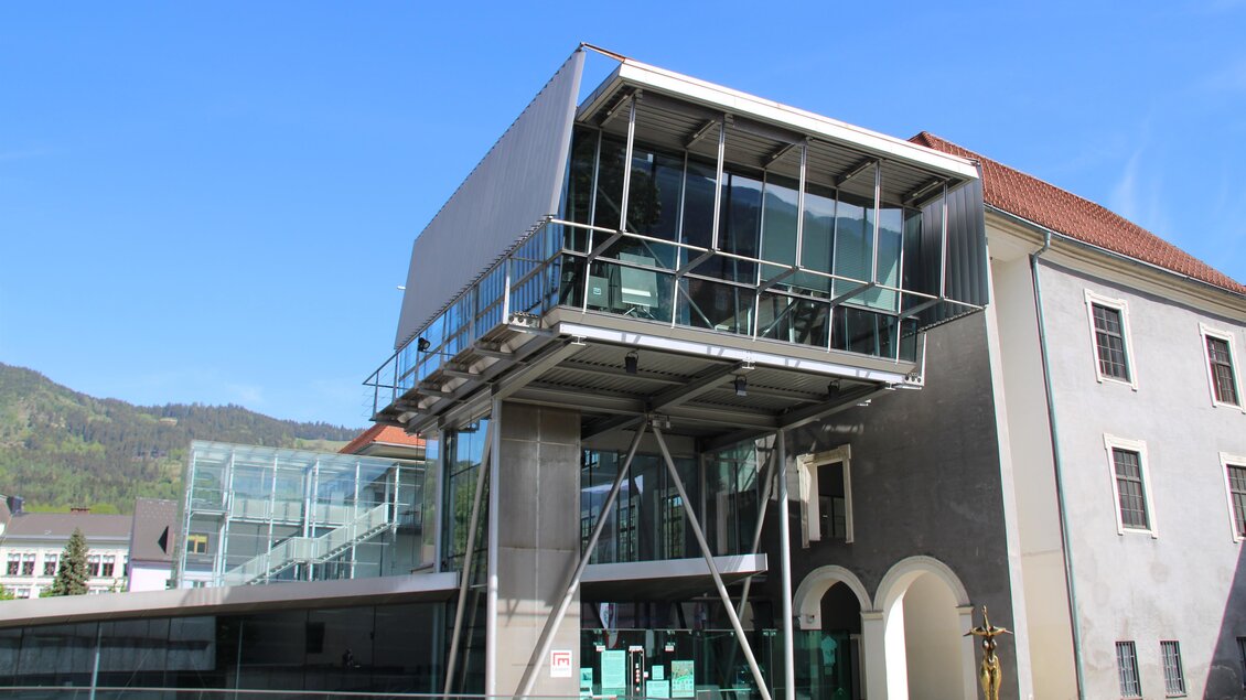 Ein modernes Gebäude mit Glasfassade und einem markanten, überhängenden Design. Es steht in einem bürgerlichen Umfeld und kombiniert Alt und Neu. | © @erzbergleoben