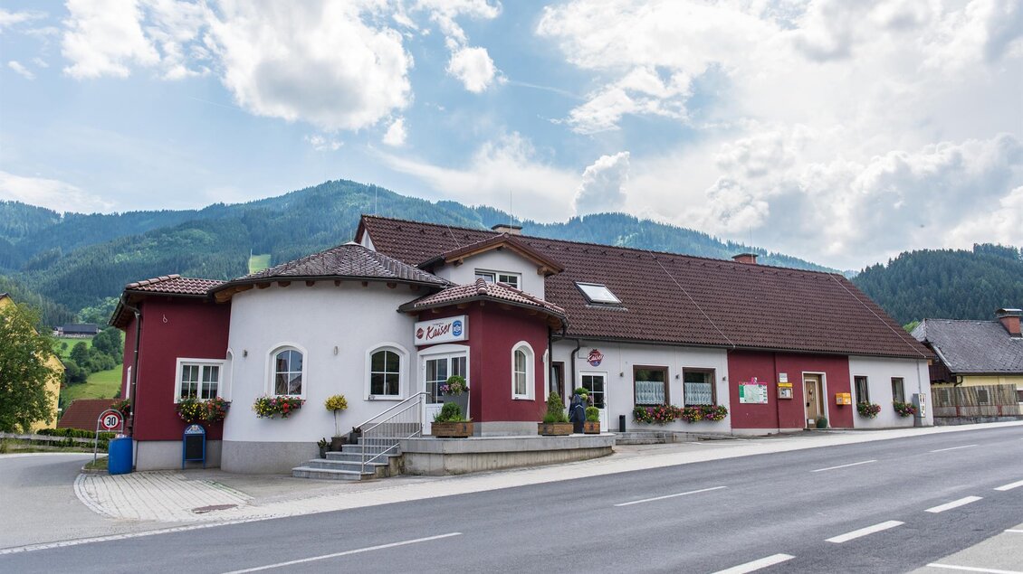 Ein charmantes Gebäude an einer Straße, umgeben von Hügeln und einem bewölkten Himmel. Das Gebäude hat einen roten und weißen Anstrich mit Blumenkästen an den Fenstern. | © Landgasthaus Kaiser