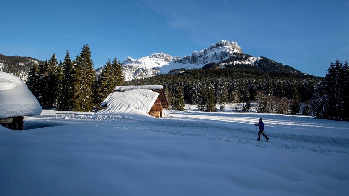 Eine winterliche Landschaft mit schneebedeckten Hütten und Tannenbäumen. Im Hintergrund sind Berge unter klarem blauen Himmel zu sehen, während eine Person im Schnee spaziert. | © TVB Ausseerland - Salzkammergut-Tom Lamm