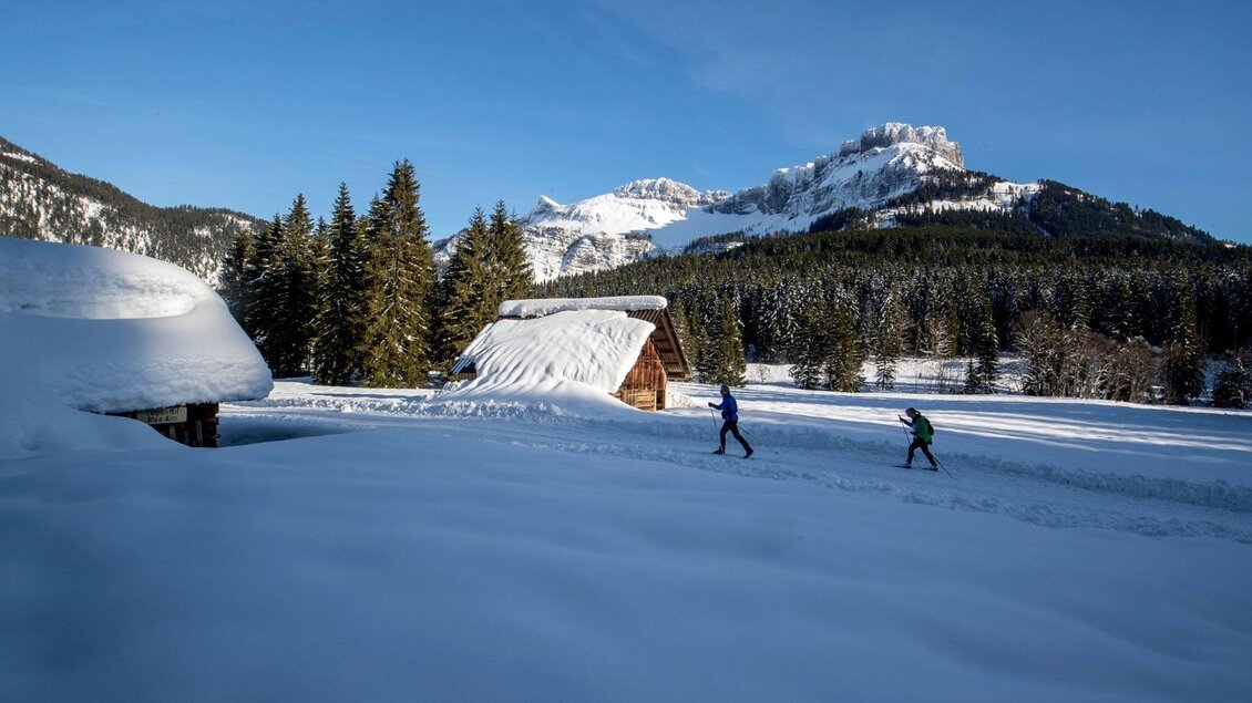 Schneebedeckte Landschaft mit Hütten und hohen Bergen im Hintergrund. Zwei Personen gehen über den verschneiten Weg. | © TVB Ausseerland - Salzkammergut-Tom Lamm