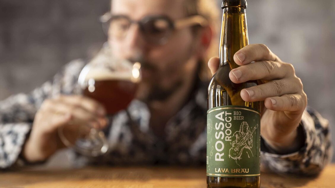Eine Person trinkt ein Glas Bier und hält eine Flasche mit dem Etikett "ROSSA" in der Hand. Der Hintergrund ist verschwommen, wodurch der Fokus auf dem Getränk liegt. | © Lava Bräu