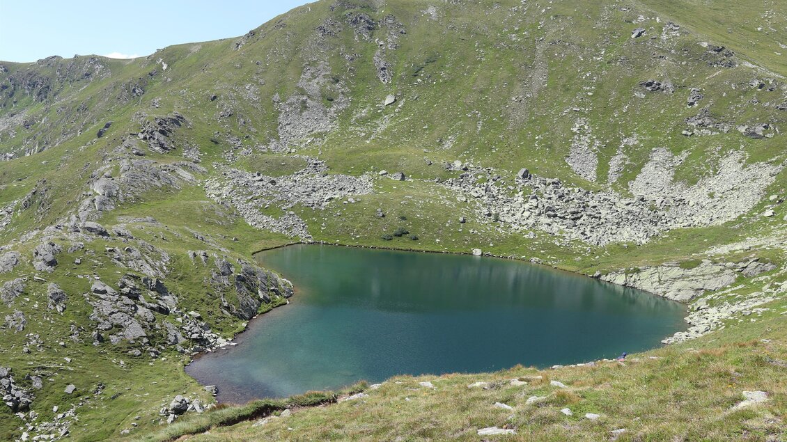 Ein klarer Bergsee umgeben von grünen Wiesen und steilen Hängen. Die Landschaft ist majestätisch und friedlich. | © WEGES