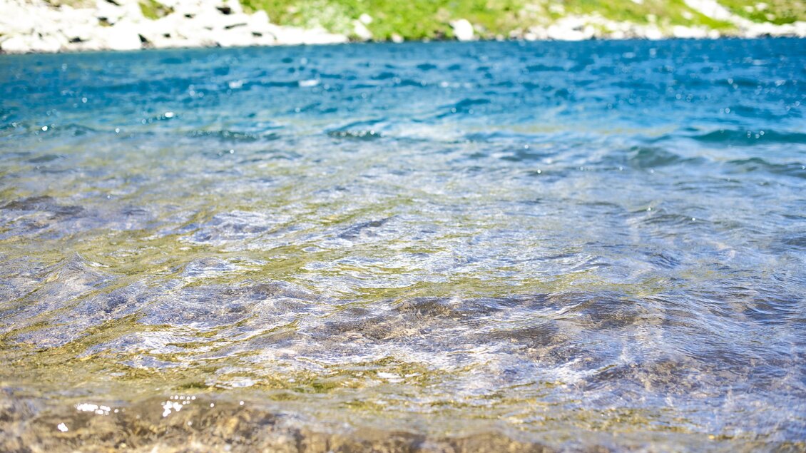 Ein klarer See mit schimmerndem Wasser in Blau- und Grüntönen. Im Hintergrund sind sanfte, grüne Hügel sichtbar. | © Anita Fössl