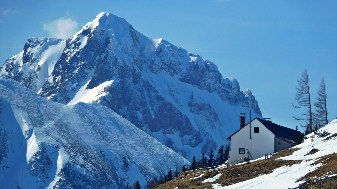 Eine schneebedeckte Berglandschaft mit einem kleinen, weißen Haus. Im Hintergrund erheben sich hohe, beeindruckende Berge unter einem klaren blauen Himmel. | © TV ERZBERG LEOBEN
