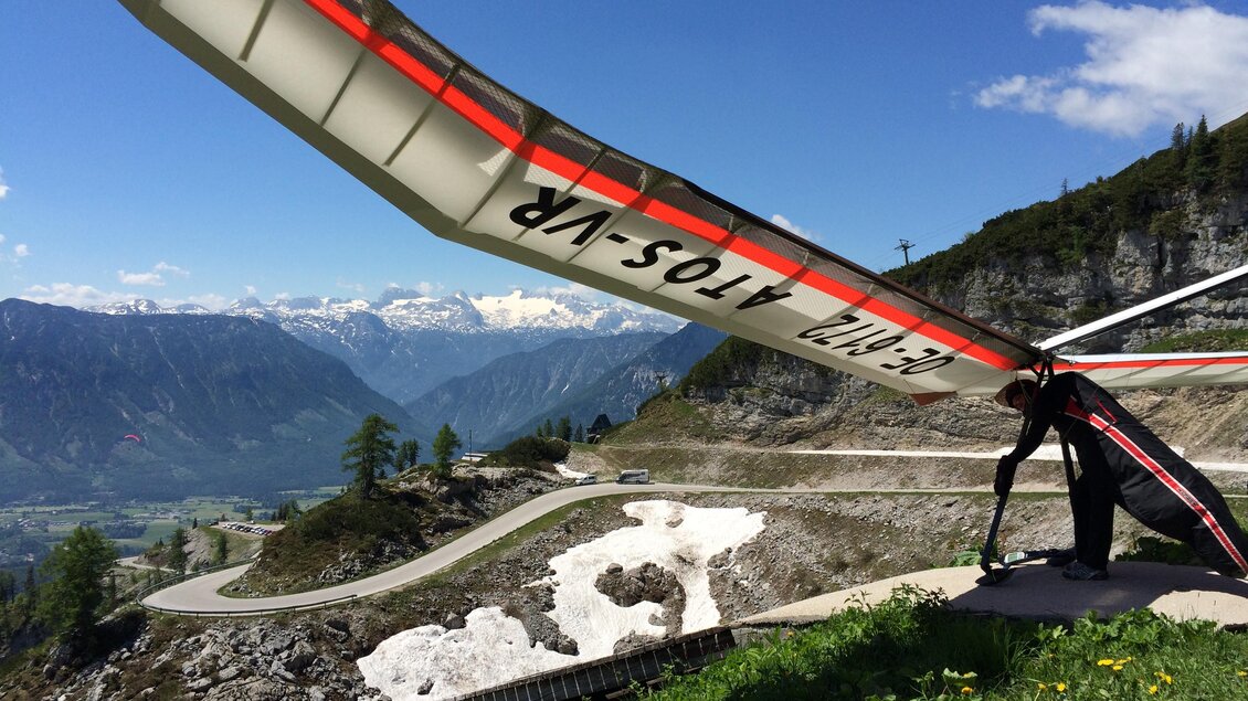 Ein Gleitschirmflieger bereitet sich auf den Abflug vom Loser in Altaussee vor. Die Landschaft mit Bergen und Almwiesen unter blauem Himmel ist atemberaubend. Im Hintergrund ist der schneebedeckte Dachstein zu sehen. | © Loser Bergbahnen/M. Raich