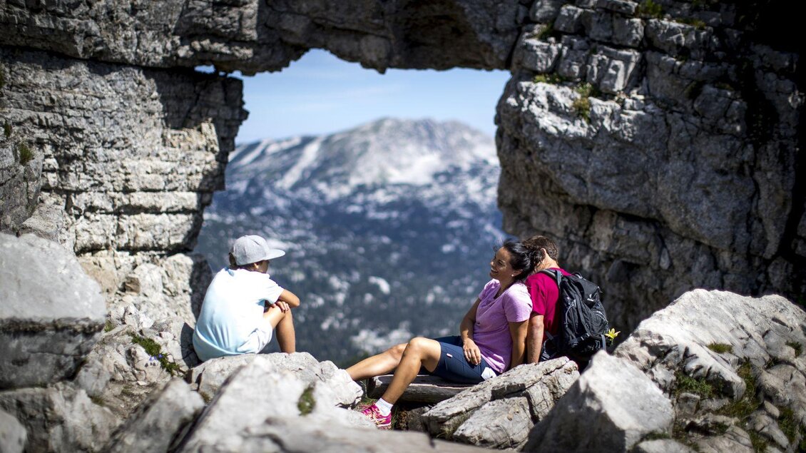 Drei Personen sitzen im Gebirge und genießen die Aussicht durch einen natürlichen Felsbogen. Die Landschaft ist beeindruckend und die Stimmung entspannt. | © TVB Ausseerland - Salzkammergut-Tom Lamm