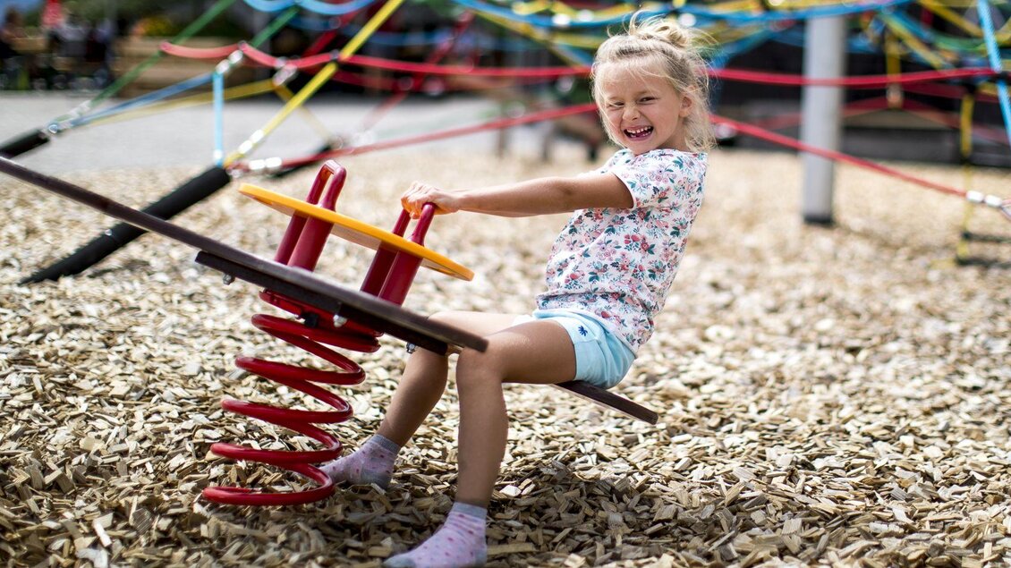 Ein fröhliches Mädchen sitzt auf einem Wippgerät auf einem Spielplatz. Der Boden ist mit Holzspänen bedeckt und bunte Bänder sind im Hintergrund zu sehen. | © TVB Ausseerland - Salzkammergut-Tom Lamm