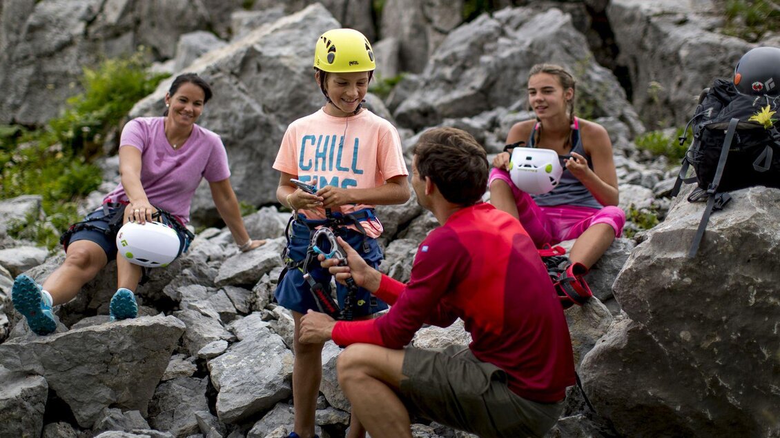 Eine Gruppe von vier Personen sitzt auf Felsen und bereitet sich auf das Klettern vor. Sie tragen Helme und haben Kletterausrüstung dabei. | © TVB Ausseerland - Salzkammergut-Tom Lamm