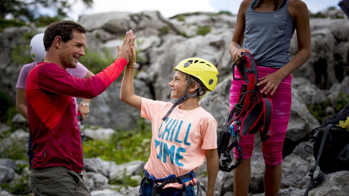 Ein Erwachsener und ein Kind geben sich ein High-Five in der Natur. Im Hintergrund sind Kletterausrüstungen und Felsen zu sehen. | © TVB Ausseerland - Salzkammergut