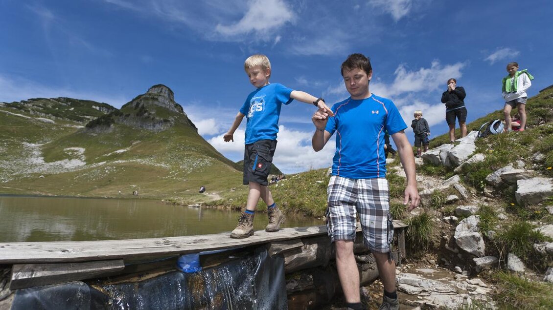Ein Vater hilft seinem Sohn, über eine kleine Brücke zu gehen. Im Hintergrund sind Berge und eine Gruppe von Menschen zu sehen. | © Loser Bergbahnen Altaussee/Leo Himsl