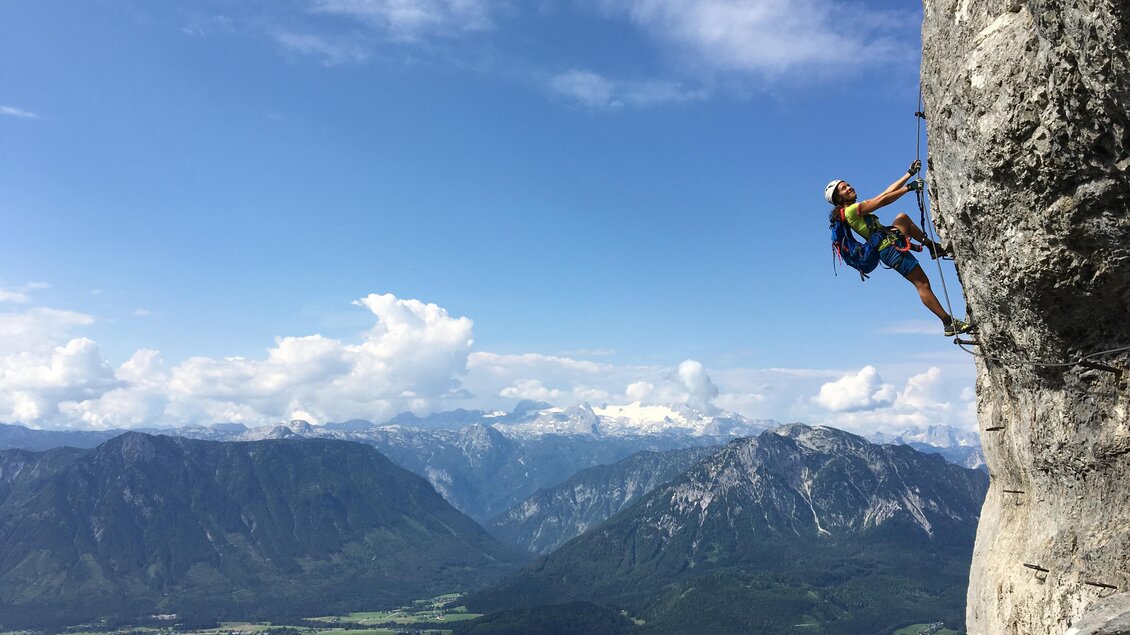 Ein Kletterer besteigt eine Felswand mit einer beeindruckenden Aussicht auf eine grüne Tallandschaft und die Berge. Der Himmel ist blau und mit einigen Wolken durchzogen. | © Loser Bergbahnen Altaussee/M. Raich