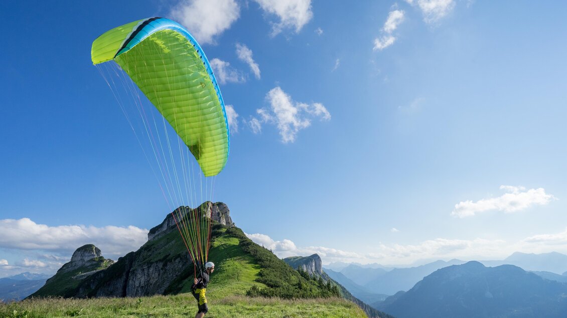 Ein Gleitschirmpilot steht auf einer grünen Wiese und bereitet sich zum Abflug vor. Im Hintergrund sind beeindruckende Berggipfel und ein blauer Himmel zu sehen. | © Loser Bergbahnen Altaussee/F. Loitzl