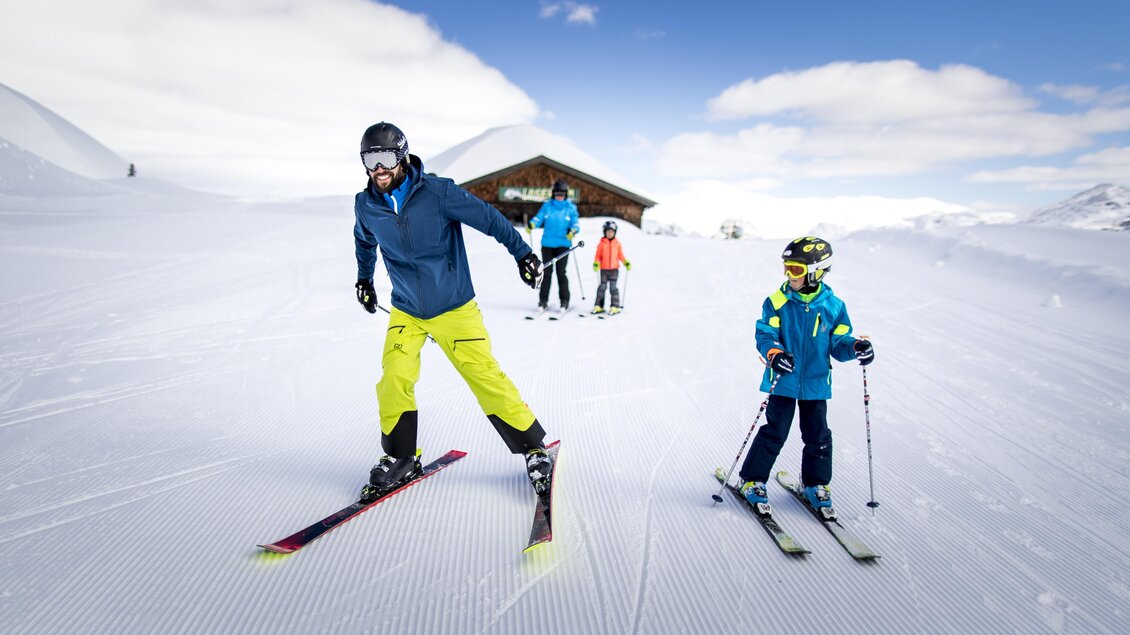 Ein Skifahrer mit leuchtend gelber Hose fährt mit einem Kind auf einer schneebedeckten Piste. Im Hintergrund sind weitere Skifahrer und ein einfaches Holzhaus zu sehen. | © TVB Ausseerland - Salzkammergut/Tom Lamm