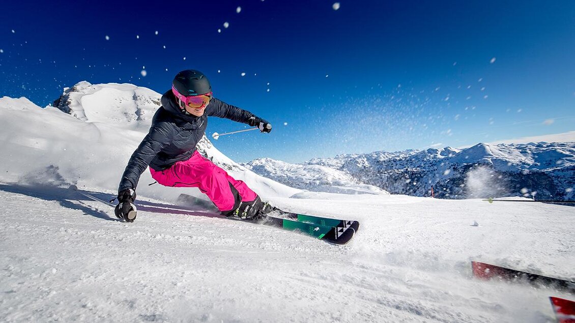 Ein Skifahrer mit einem pinken Skihosen fährt auf frischem Schnee. Im Hintergrund sind majestätische Berge und ein blauer Himmel zu sehen. | © TVB Ausseerland - Salzkammergut / Tom Lamm