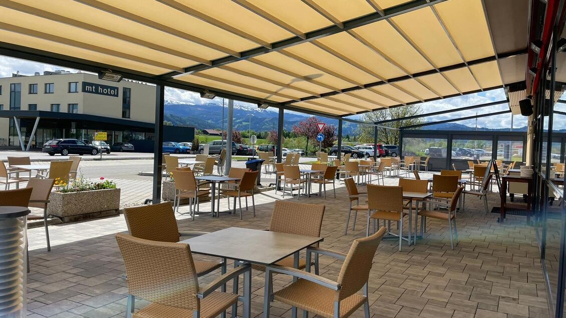Eine gemütliche Terrasse mit Tischen und Stühlen unter einem Sonnenschutz. Im Hintergrund sind Bäume und Gebäude zu sehen. | © M-Rast