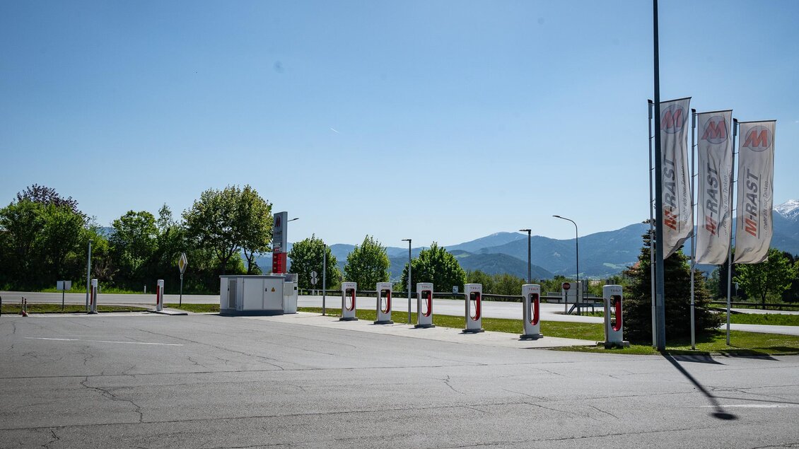 Ein leerer Parkplatz mit einigen Tankstellen. Im Hintergrund sind grüne Bäume und Berge zu sehen. | © M-Rast