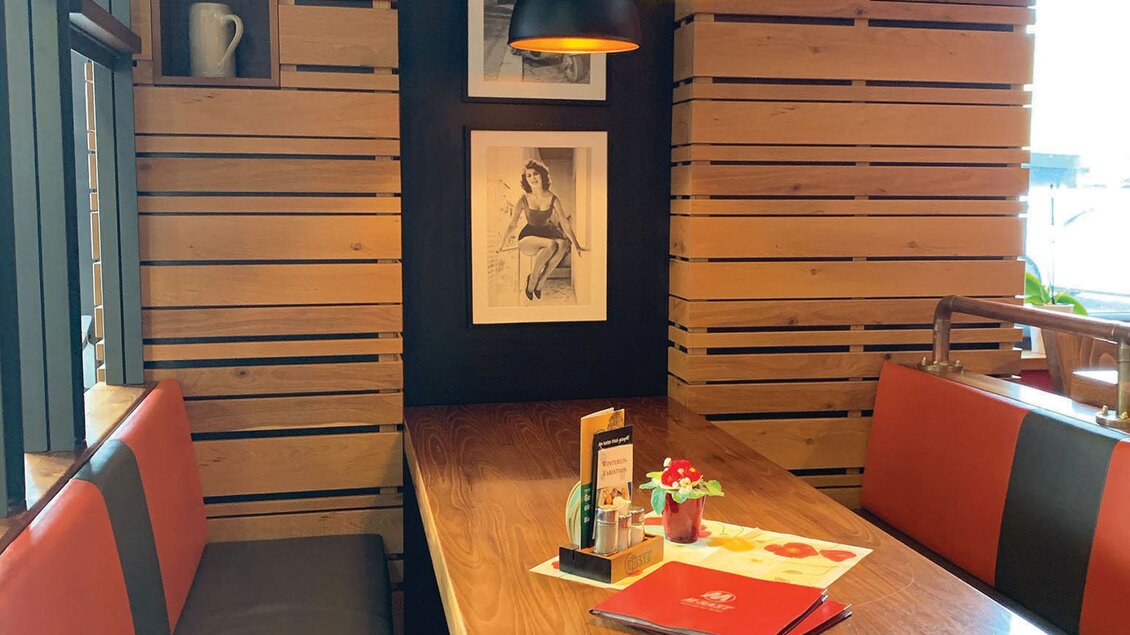 Ein gemütlicher Restaurantbereich mit einer Holzwand und einer Tischdekoration. Die Beleuchtung ist modern und die Sitzbänke sind rot und schwarz. | © M-Rast