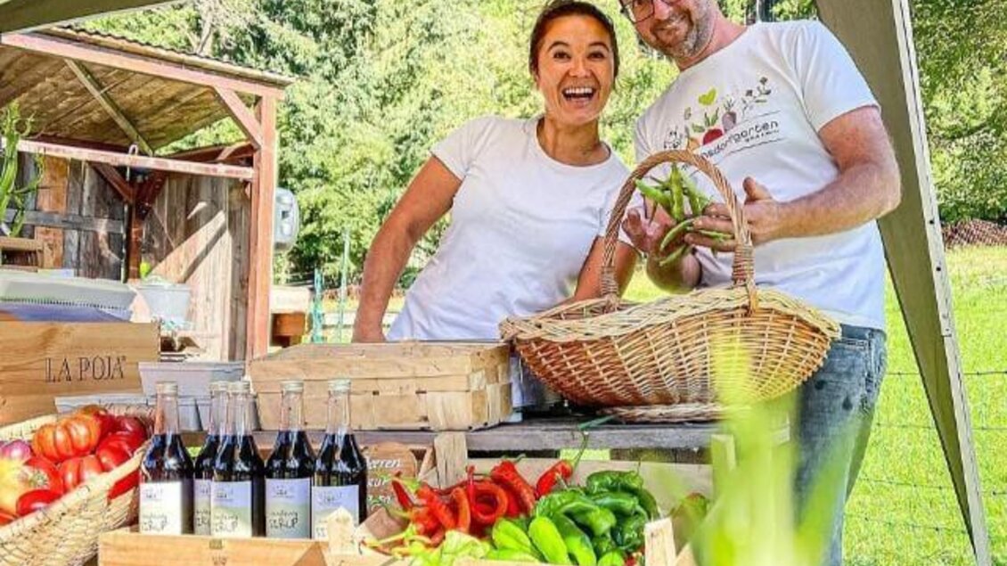 Ein freundliches Paar steht an einem Marktstand. Sie präsentieren frisches Gemüse und Getränke unter einem Sonnenschirm. | © Villmannsdorfgarten