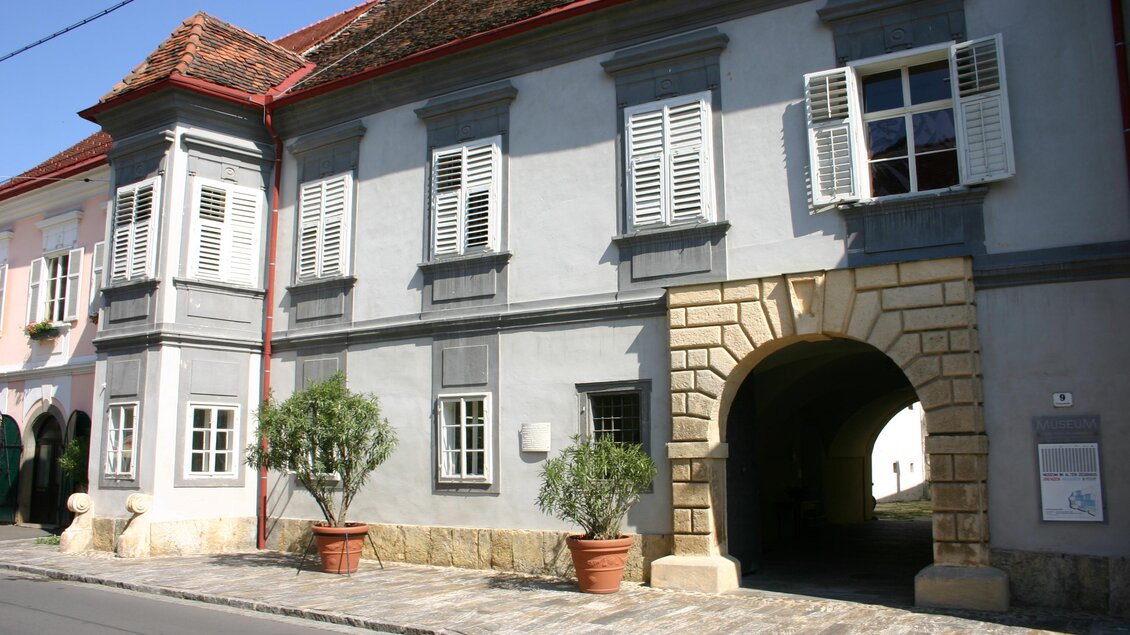 Ein historisches Gebäude mit grauer Fassade und großen Fenstern. Es hat einen Bogen im Eingangsbereich und ist von Pflanzen in Töpfen umgeben. | © MiaZ