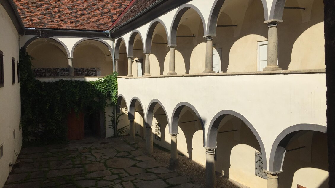 Ein historischer Innenhof mit eleganten Bögen. Der Boden besteht aus großen Steinen, und grüne Pflanzen wachsen an der Wand. | © MiaZ