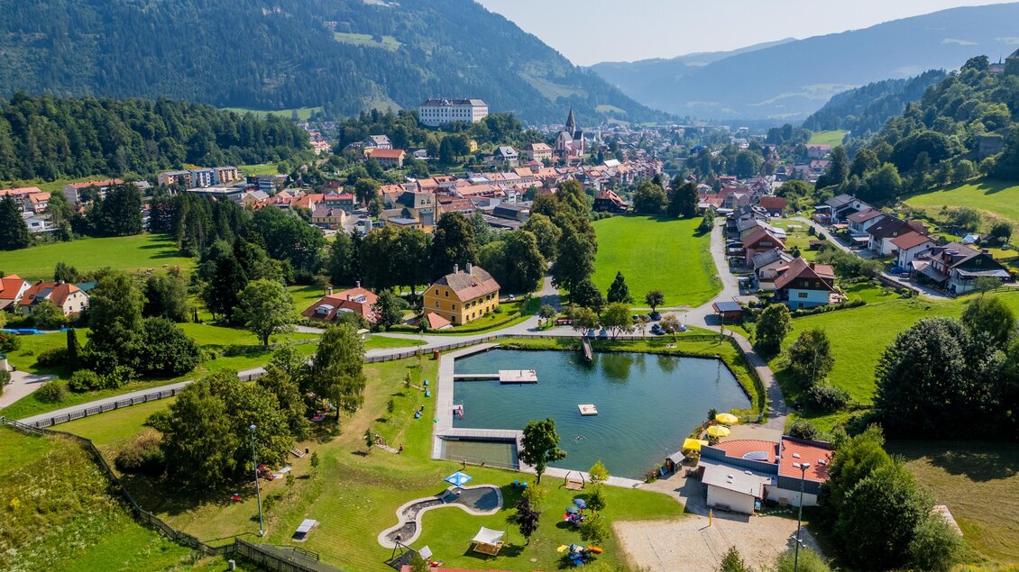 Eine malerische Landschaft mit einem ruhigen See und umliegenden Wiesen. Im Hintergrund ist ein kleines Dorf mit grünen Hügeln und Bergen zu sehen. | © TV Murau