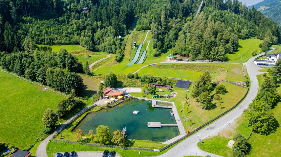 Eine malerische Landschaft mit einem Teich und grünen Wiesen. Im Hintergrund sind Skihänge und eine klare blaue Himmel sichtbar. | © TV Murau