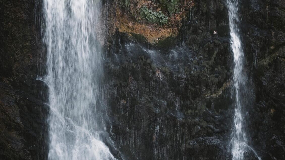 Ein Mann in einem gelben Regenmantel steht vor zwei beeindruckenden Wasserfällen. Um ihn herum ist eine natürliche, felsige Landschaft. | © Stefan Leitner
