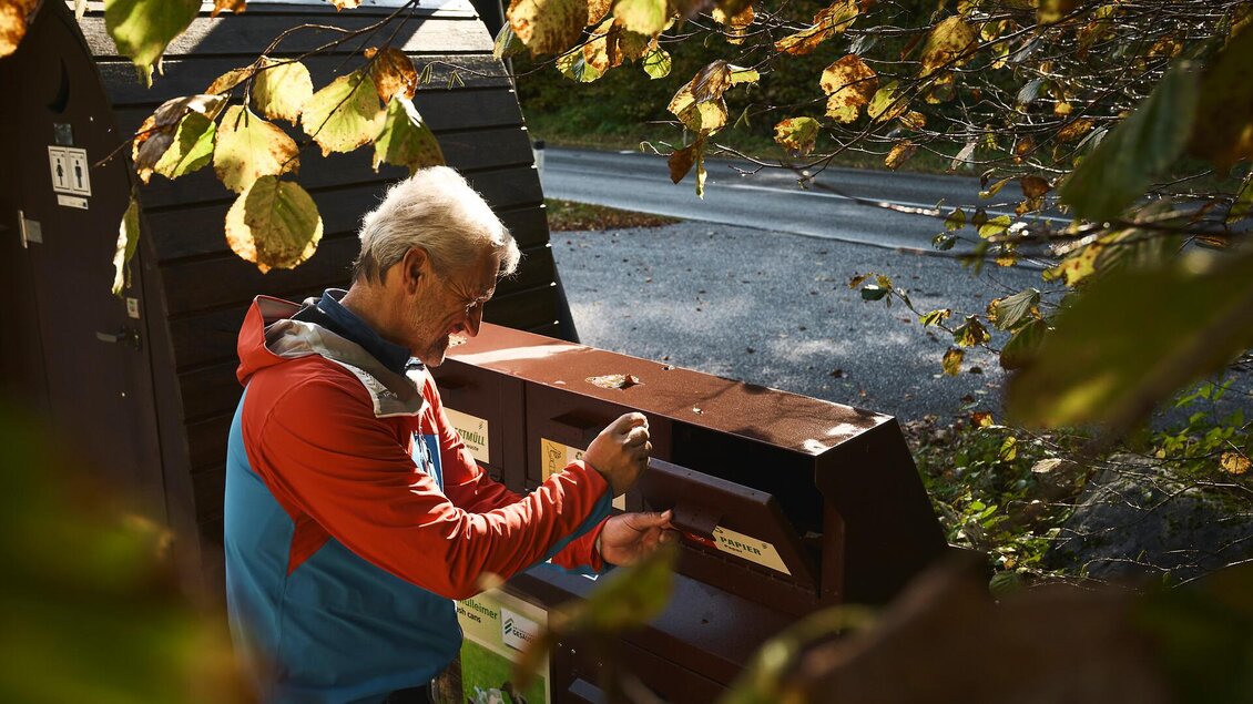Ein Mann steht an einem Recyclingcontainer und wirft etwas hinein. Die Umgebung ist von herbstlichen Blättern und einem ruhigen Weg umgeben.