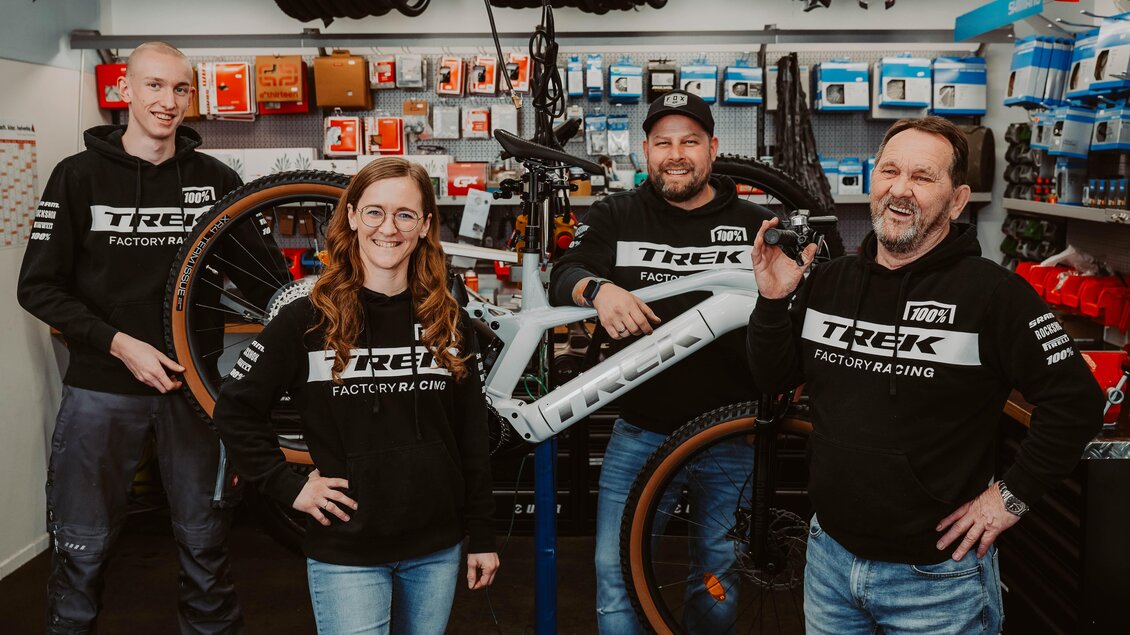 Ein Team von vier Personen steht in einem Fahrradgeschäft, umgeben von Fahrrädern und Werkzeugen. Sie tragen schwarze Hoodies mit dem Logo von Trek und lächeln in die Kamera. | © Radwerkstatt Willibald