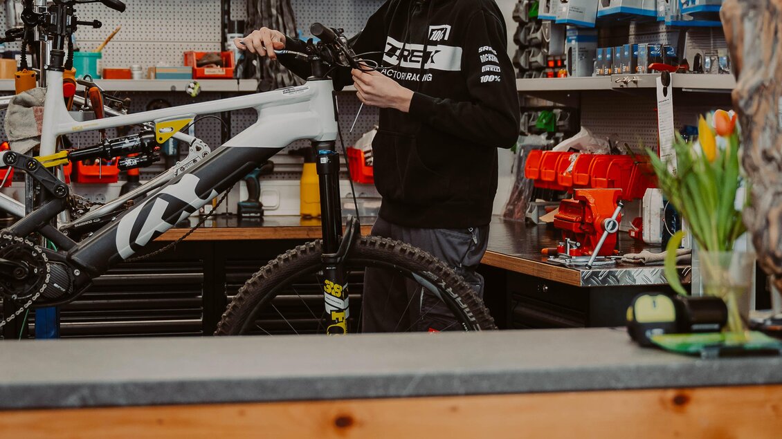 Ein Fahrradmechaniker arbeitet an einem Mountainbike in einem Fahrradgeschäft. Im Hintergrund sind verschiedene Fahrradteile und Zubehörteile sichtbar. | © Radwerkstatt Willibald