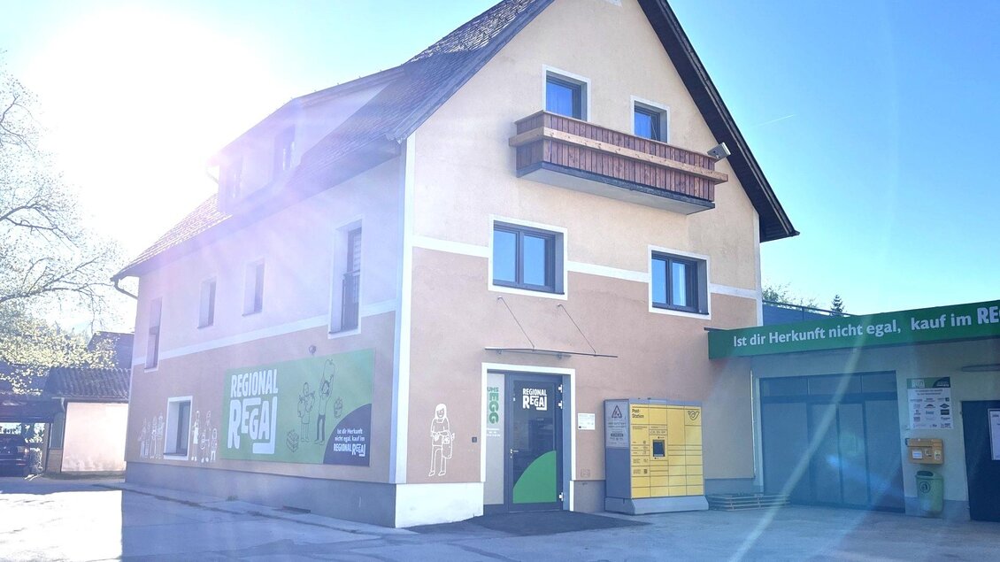 Ein beige-farbiges Gebäude mit einem Balkon und mehreren Fenstern. Im Vordergrund sind Parkplätze und ein gelbes Postfach sichtbar.