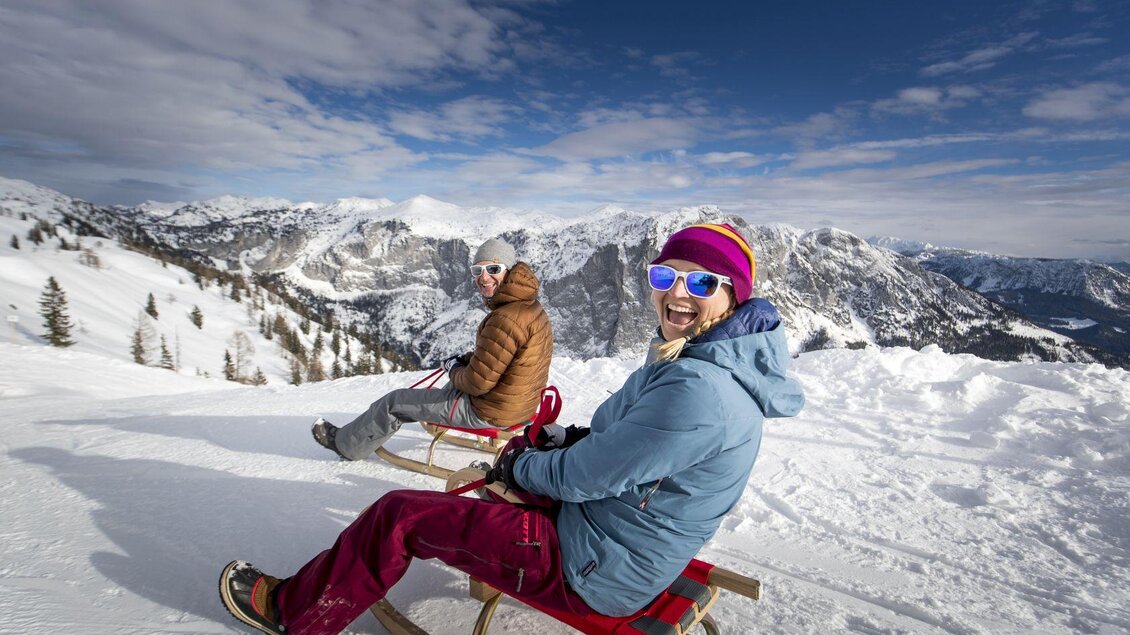 Zwei Menschen sitzen auf Schlitten im Schnee und lachen. Im Hintergrund sind schneebedeckte Berge und ein blauer Himmel zu sehen. | © TVB Ausseerland - Salzkammergut-Tom Lamm