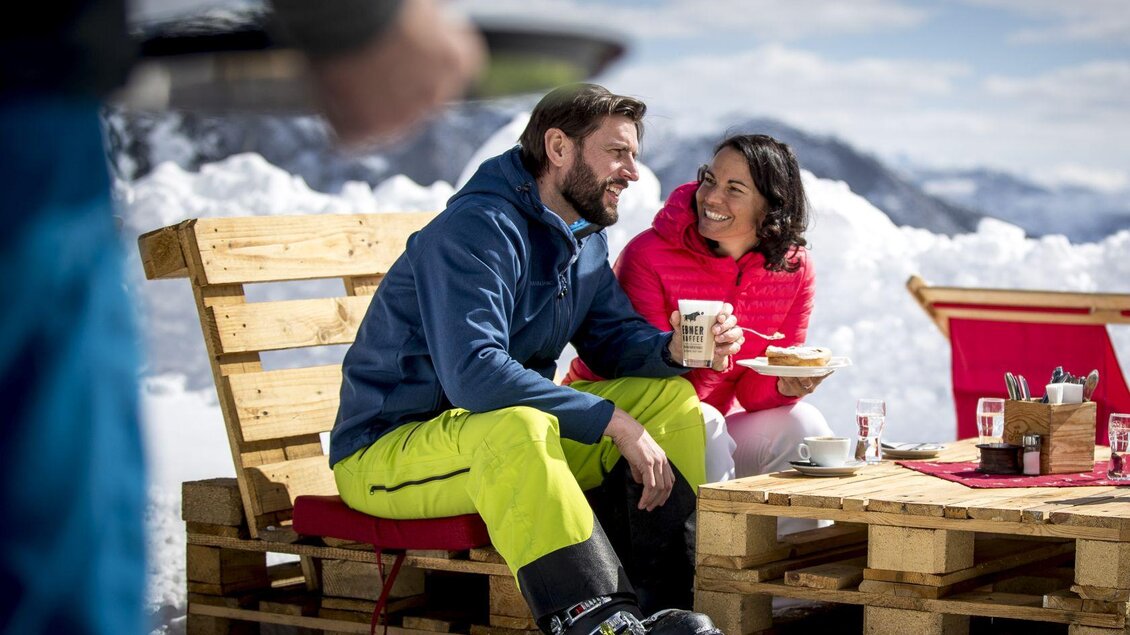 Ein Paar genießt eine Pause in den Bergen. Sie sitzen auf Holzmöbeln im Schnee und lachen miteinander. | © TVB Ausseerland - Salzkammergut-Tom Lamm