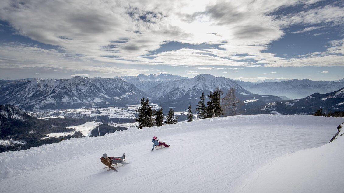 Eine verschneite Landschaft mit Bergen im Hintergrund. Zwei Rodelfahrer genießen die Abfahrt auf der Piste. | © TVB Ausseerland - Salzkammergut-Tom Lamm