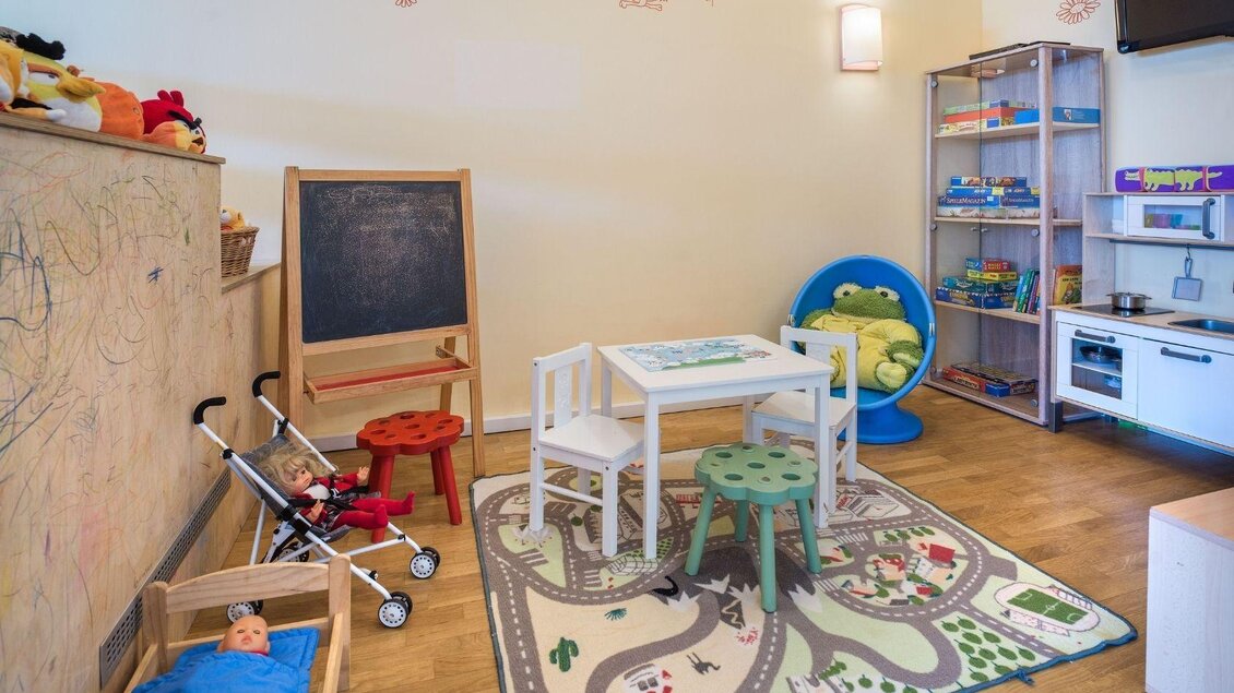 Ein farbenfroher Spielraum für Kinder mit einem Tisch, Stühlen und einem Teppich, der Straßenmuster zeigt. Es gibt Spielzeug, eine Kreidetafel und eine gemütliche Kuschelliege. | © JUFA Hotels