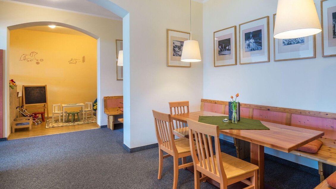 Ein gemütlicher Speisebereich mit einem Holztisch und Stühlen. An der Wand hängen Bilder, und im Hintergrund ist ein Spielbereich für Kinder zu sehen. | © JUFA Hotels