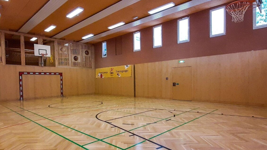 Eine Sporthalle mit einem Holzboden und einem Basketballkorb. An einer Wand befindet sich ein Handballtor und die Fenster sorgen für natürliches Licht. | © JUFA Hotels