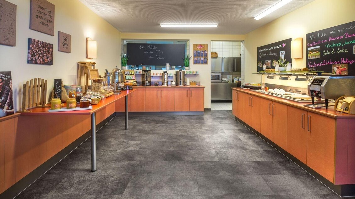 Ein einladender Frühstücksraum mit Holzregalen und einer Auswahl an Speisen. Die Wände sind mit Tafeln dekoriert, die Menüs anzeigen. | © JUFA Hotels