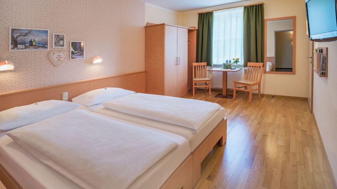 Ein gemütliches Hotelzimmer mit zwei Einzelbetten, Holzfußboden und sanften Farben. Ein Tisch mit Stühlen und ein Fenster mit Vorhängen sorgen für eine einladende Atmosphäre. | © JUFA Hotels